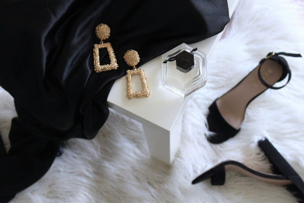 A black dress and heels beside a bottle of perfume on a soft white rug, creating an elegant fashion display.