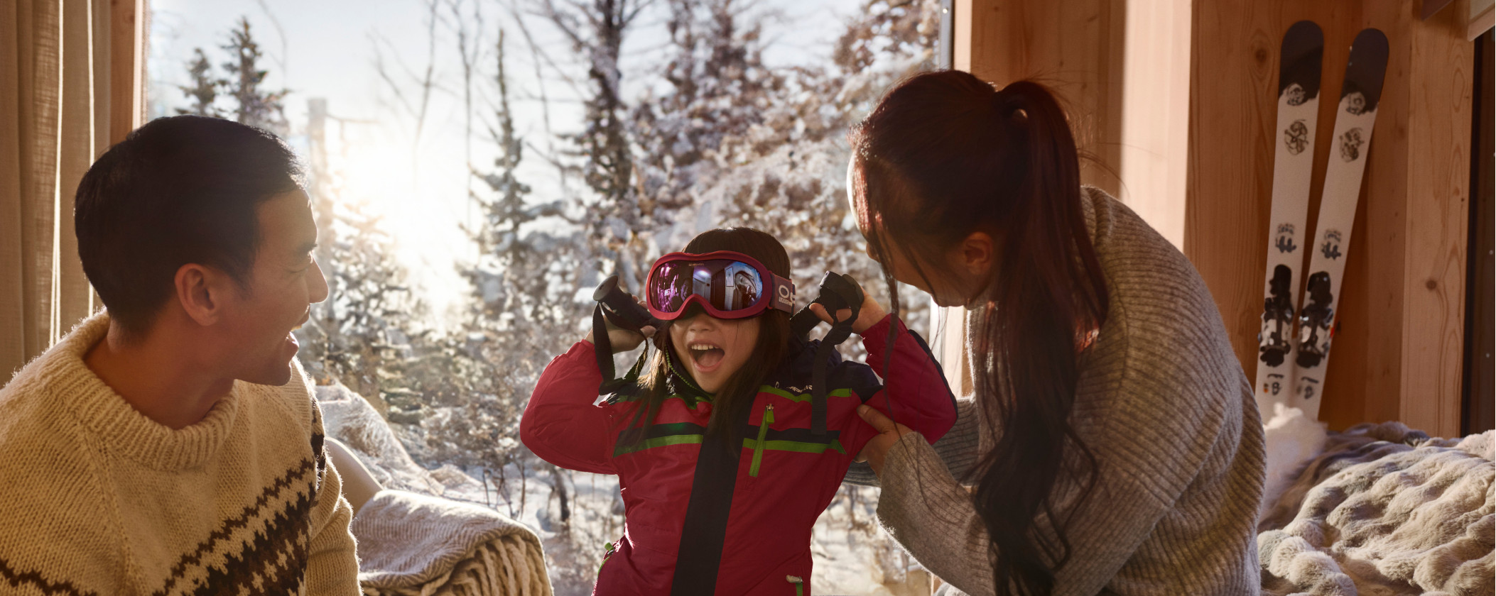 A family of three sits together in a cozy room, surrounded by skis and winter gear, smiling and enjoying each other's company.